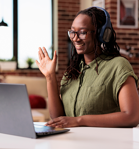 A woman with headphones on smiling and waving to the webcam on her laptop